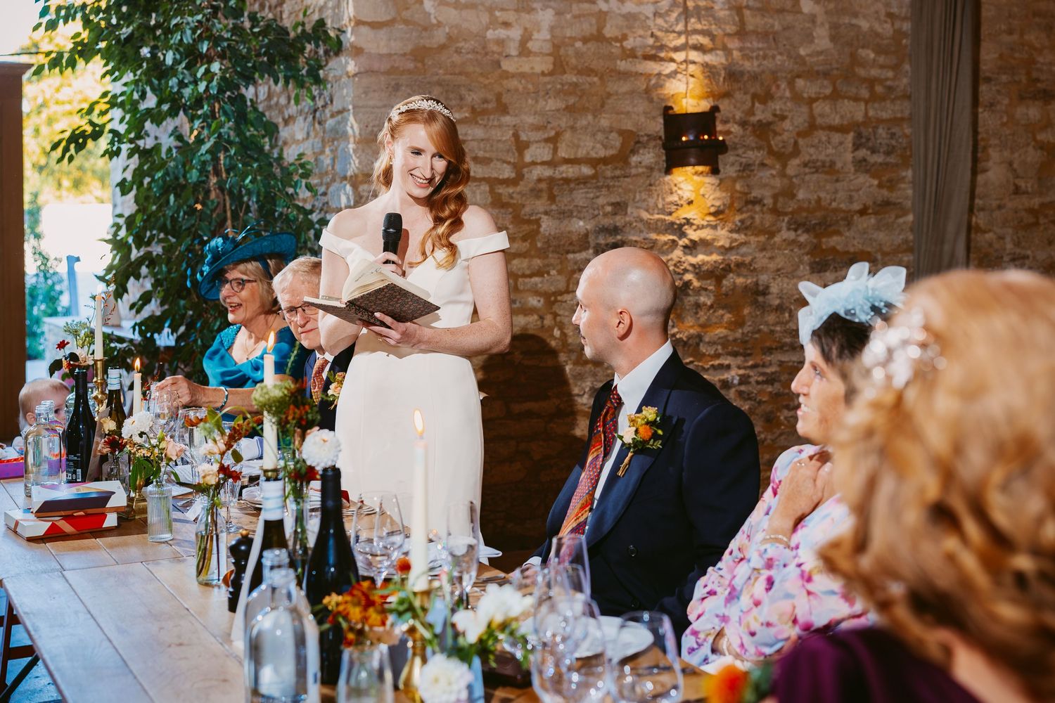 A smiling bride in a white dress reads from a notebook while giving a speech at a candlelit wedding table with seated guests.