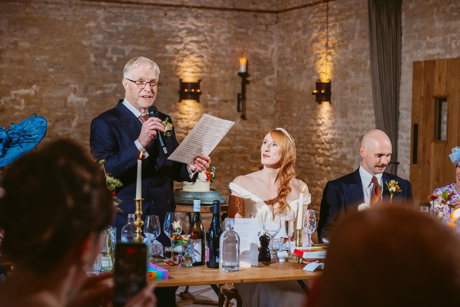 A man in a navy suit reads a wedding speech from paper at a candlelit reception table beside the bride and groom.