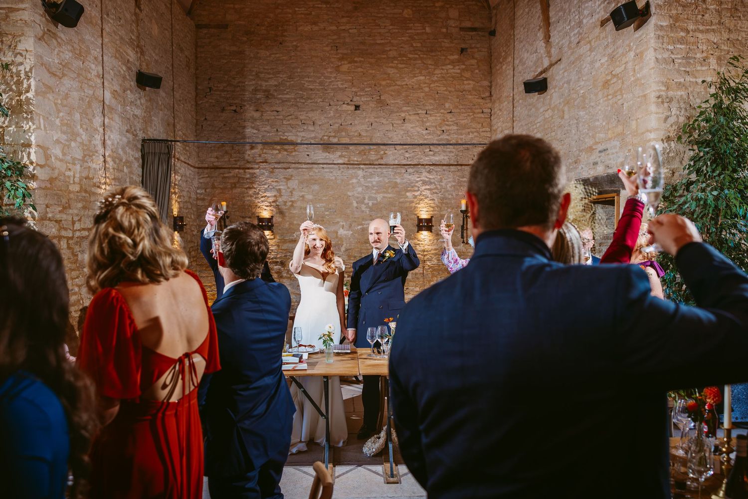 Wedding guests stand and raise their arms to cheer the bride and groom at a rustic brick venue with warm lighting and greenery.