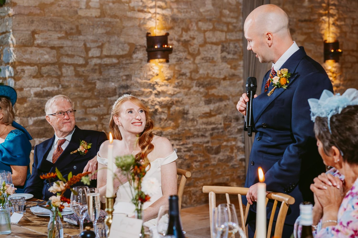Groom in a navy suit gives a wedding speech into a microphone while the smiling bride sits at a candlelit reception table nearby.
