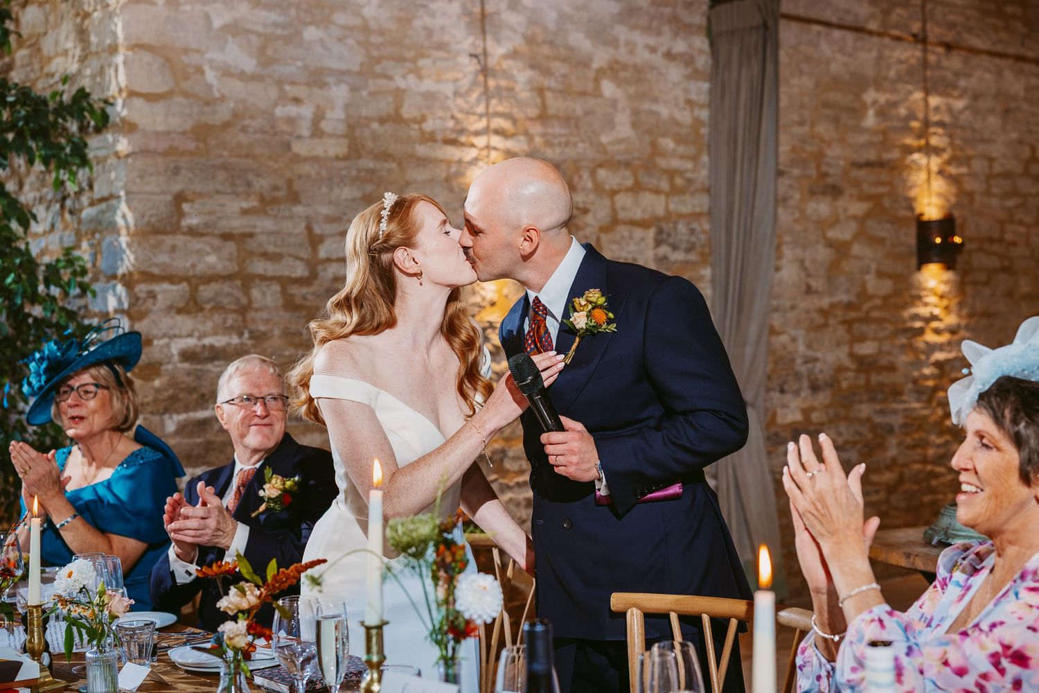 Bride and groom share a kiss during a wedding reception as smiling guests in fascinators applaud beside candlelit tables.