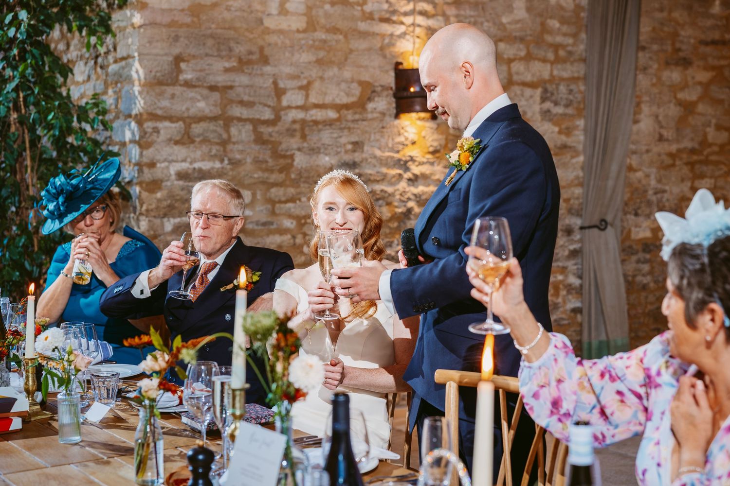 A groom in a navy suit raises a glass for a toast beside his smiling bride as wedding guests look on.