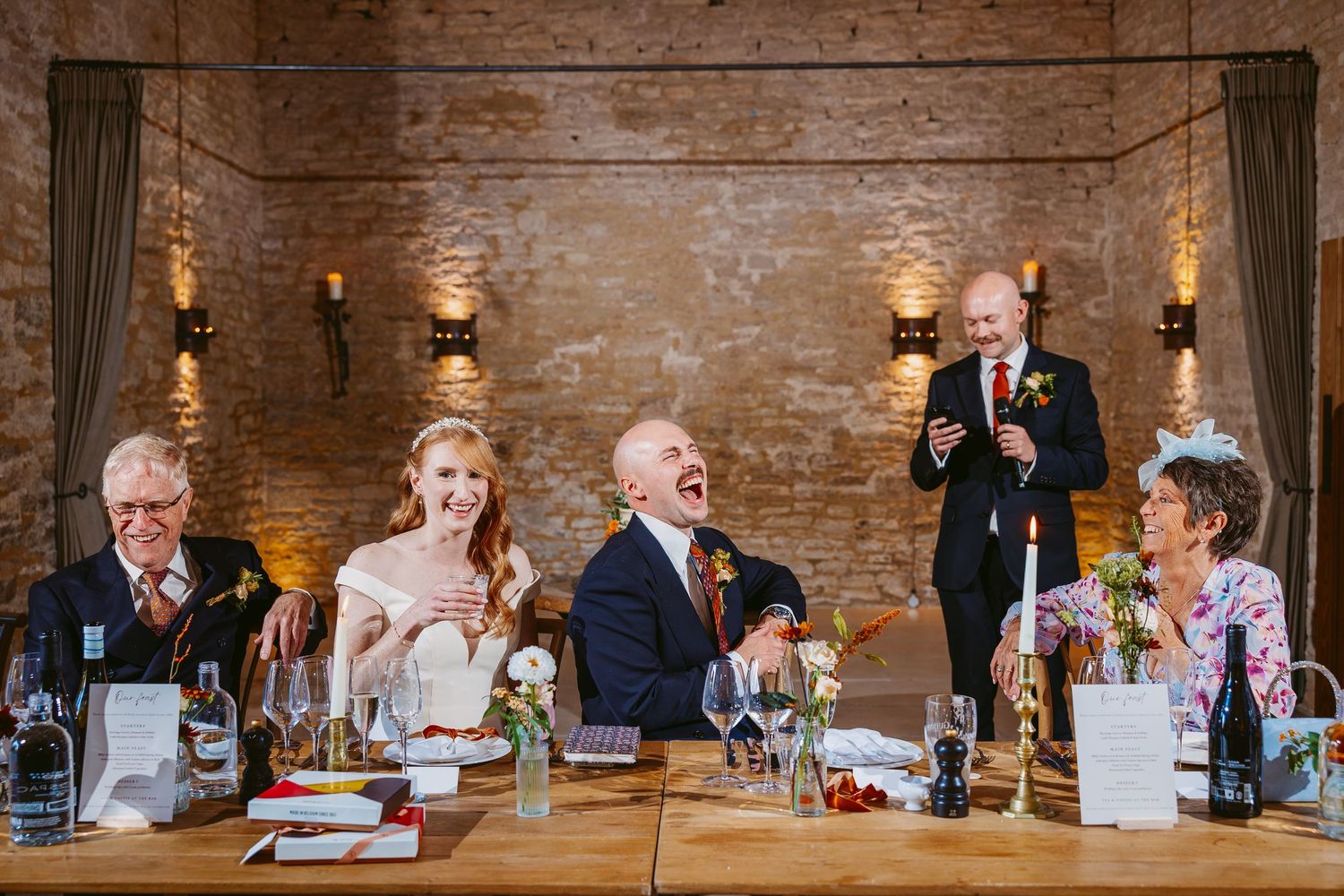 Bride, groom and wedding guests laugh at the head table during speeches in a rustic stone-walled reception hall.
