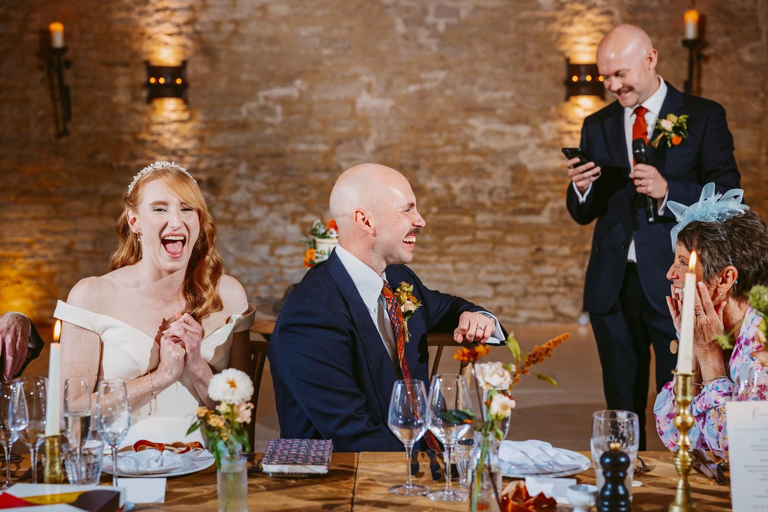 Bride laughs joyfully as a man in a suit reads his wedding speech at the reception table lit by candles.