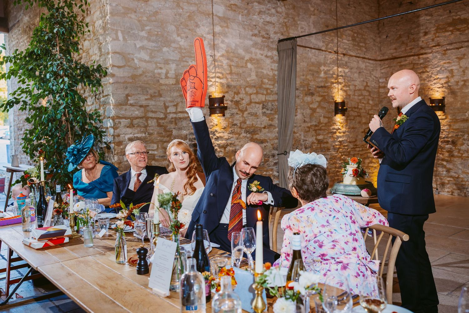 A wedding guest raises a hand holding an orange item while the groom stands with a microphone giving a speech at the reception tables.