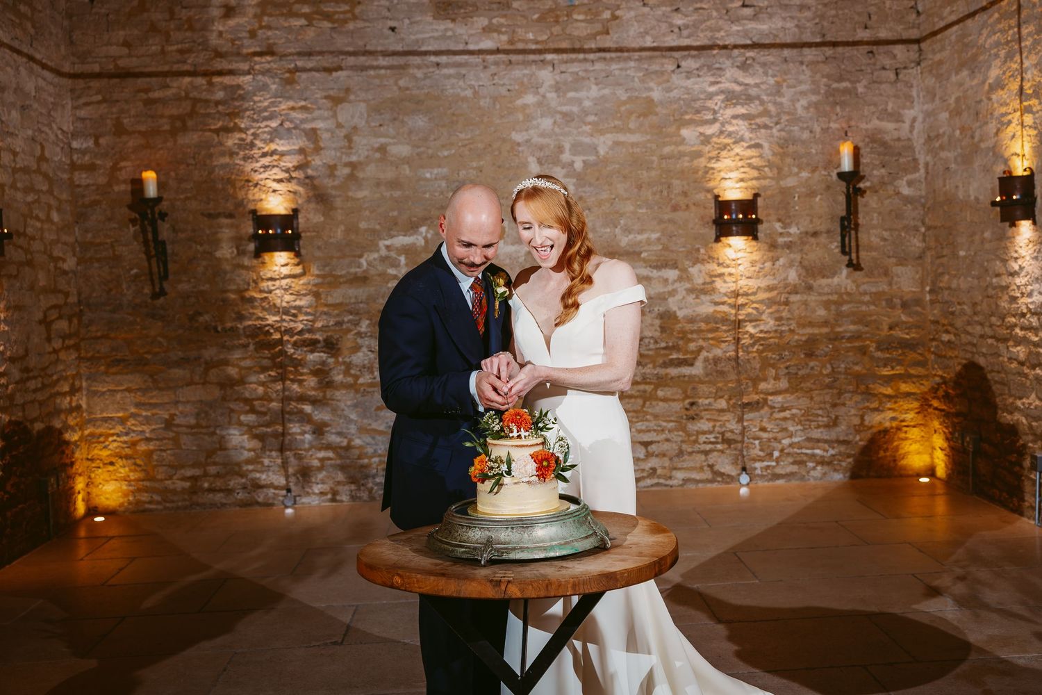 Bride and groom cut their floral-decorated tiered wedding cake together in a rustic stone-walled venue lit by warm wall sconces.
