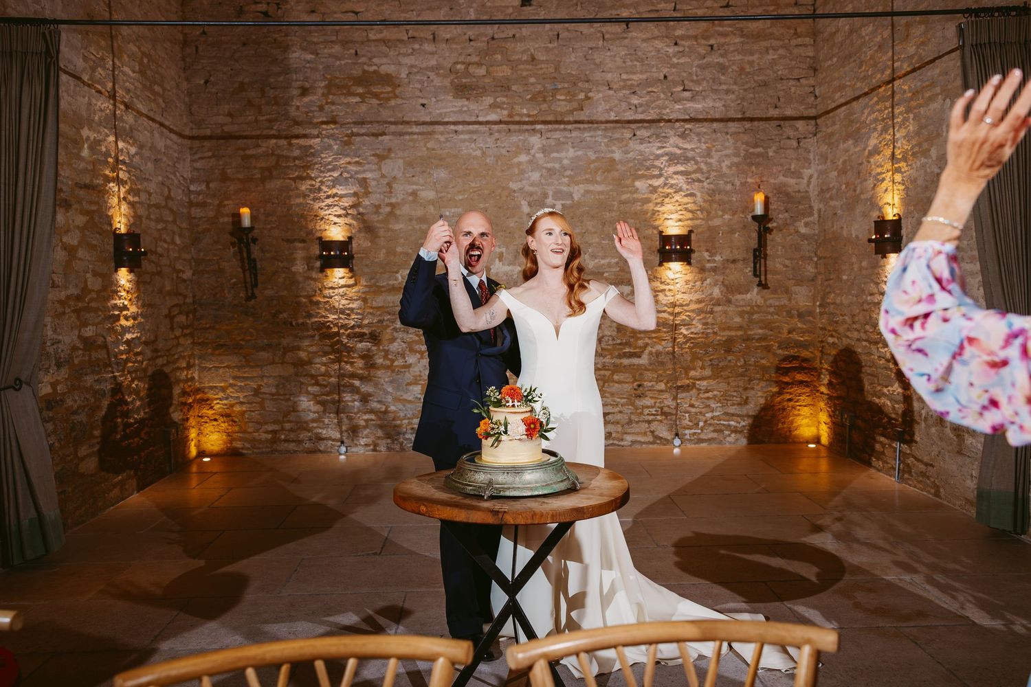Bride and groom raise their hands in celebration beside a wedding cake in a candlelit brick-walled reception room.