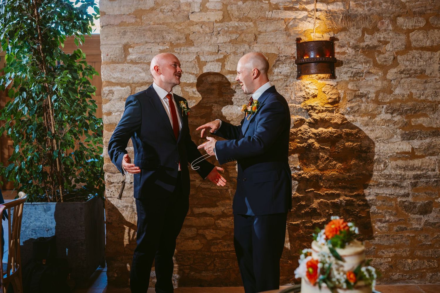 Two men in dark suits share a lighthearted moment beside a stone wall and an autumn flower arrangement at a wedding.