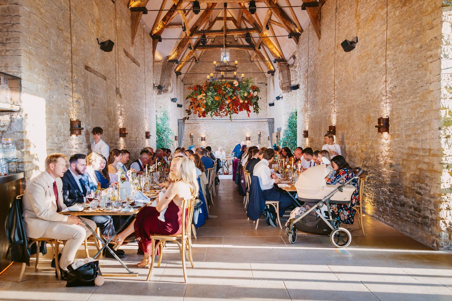 Wedding guests sit at long tables in a rustic stone barn venue with wooden beams and a floral arrangement at the far end.