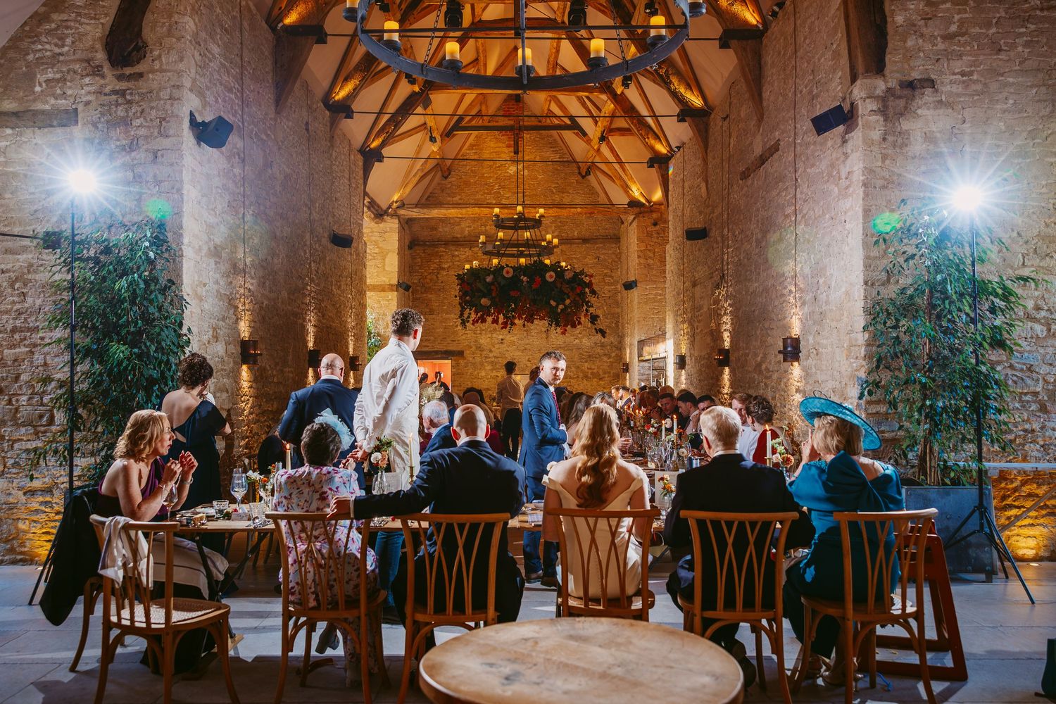 Wedding guests seated at tables in a rustic stone barn venue with exposed timber beams, chandeliers and warm uplighting during a ceremony.