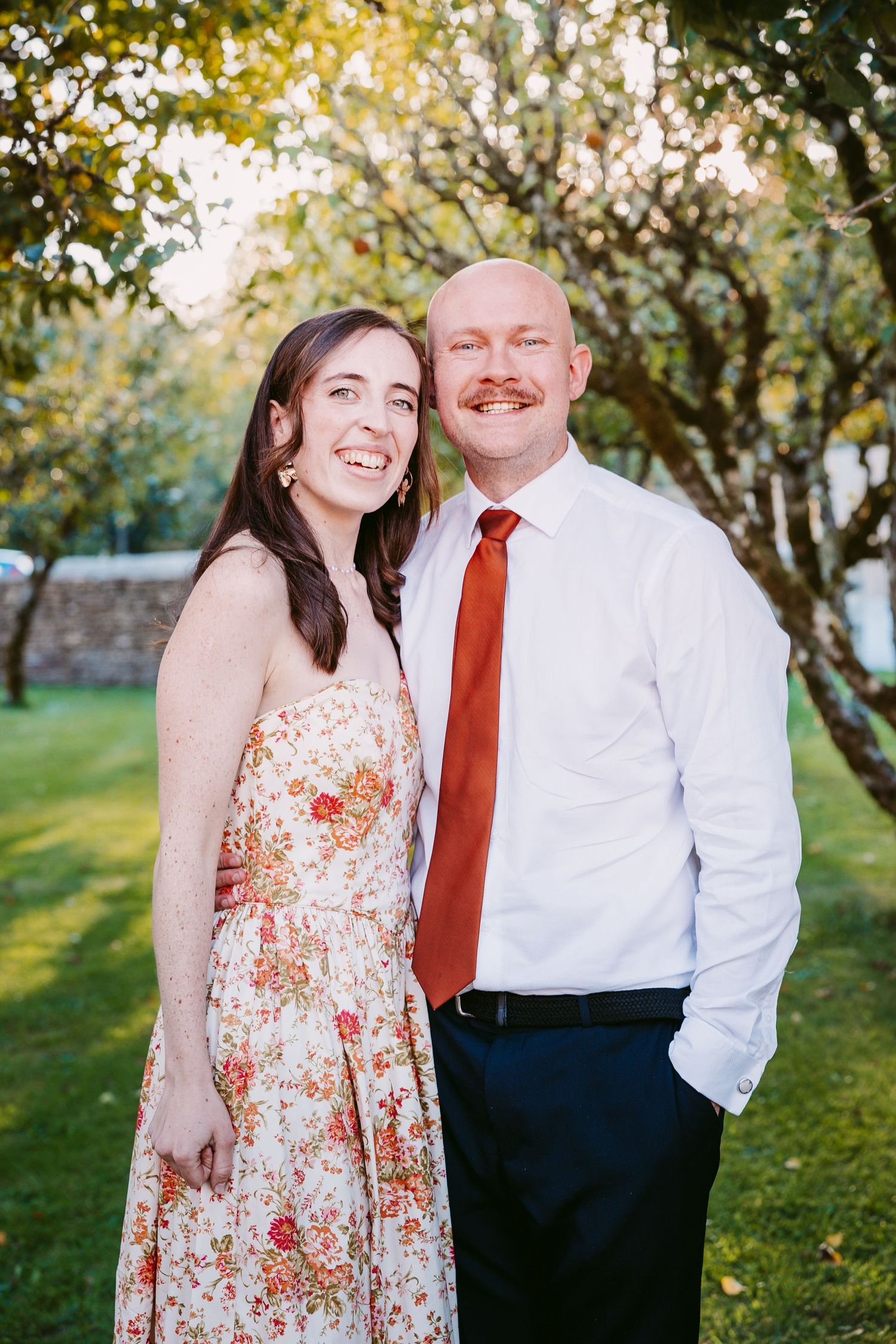 Smiling couple in a floral sundress and white shirt with rust-orange tie pose together in an orchard on a sunny day.