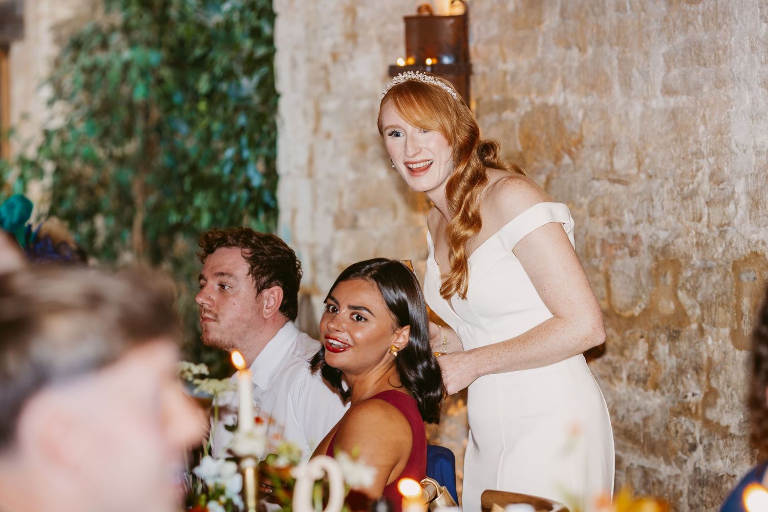 Red-haired bride in a white dress stands smiling at a candlelit wedding reception table beside seated guests.