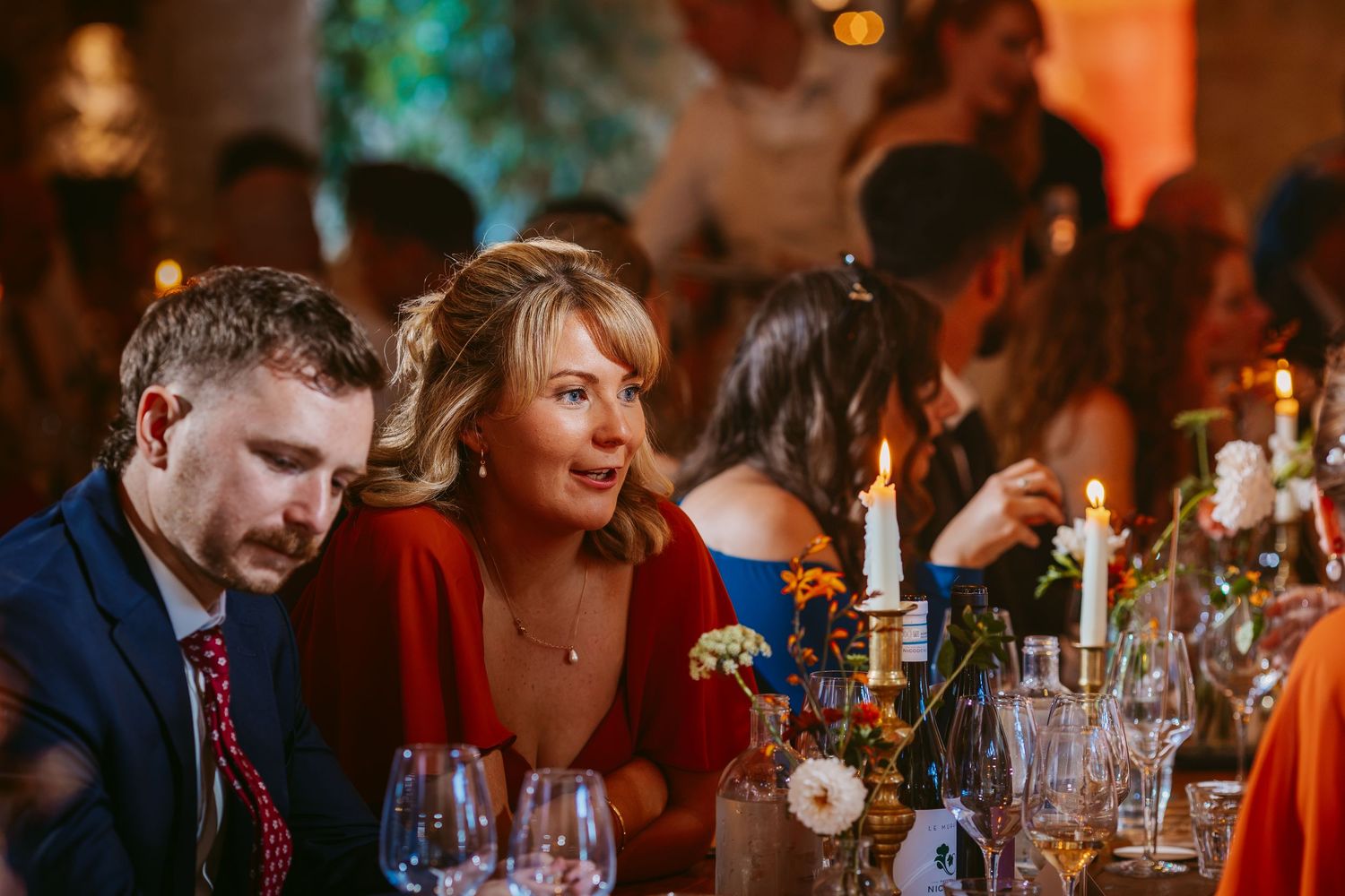 Smiling woman in a red dress and a man in a navy suit sit at a candlelit wedding reception table with flowers and glassware.