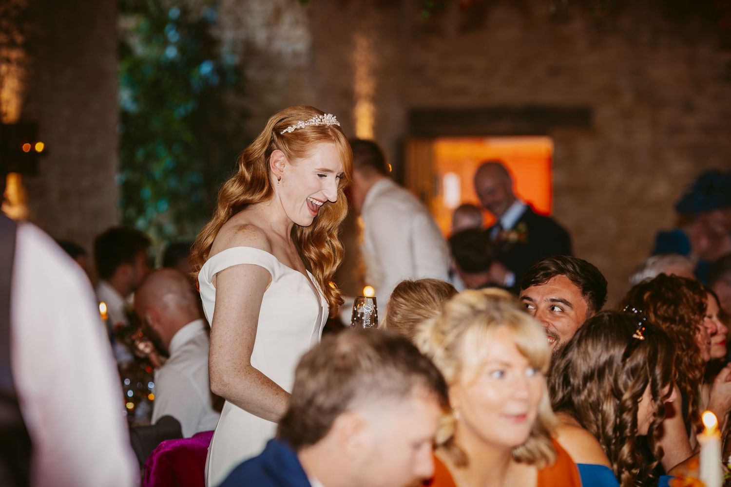 Smiling bride in a white dress and floral hair accessory stands among seated wedding guests at a candlelit reception in a rustic brick venue