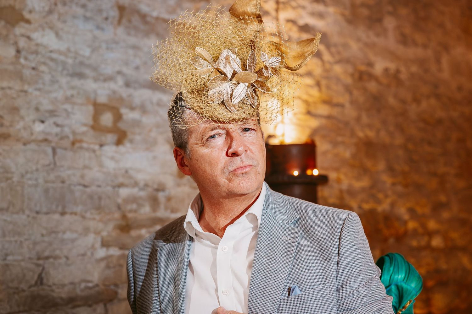A man in a gray blazer wears a gold feathered fascinator at a wedding reception inside a warm, rustic stone venue.