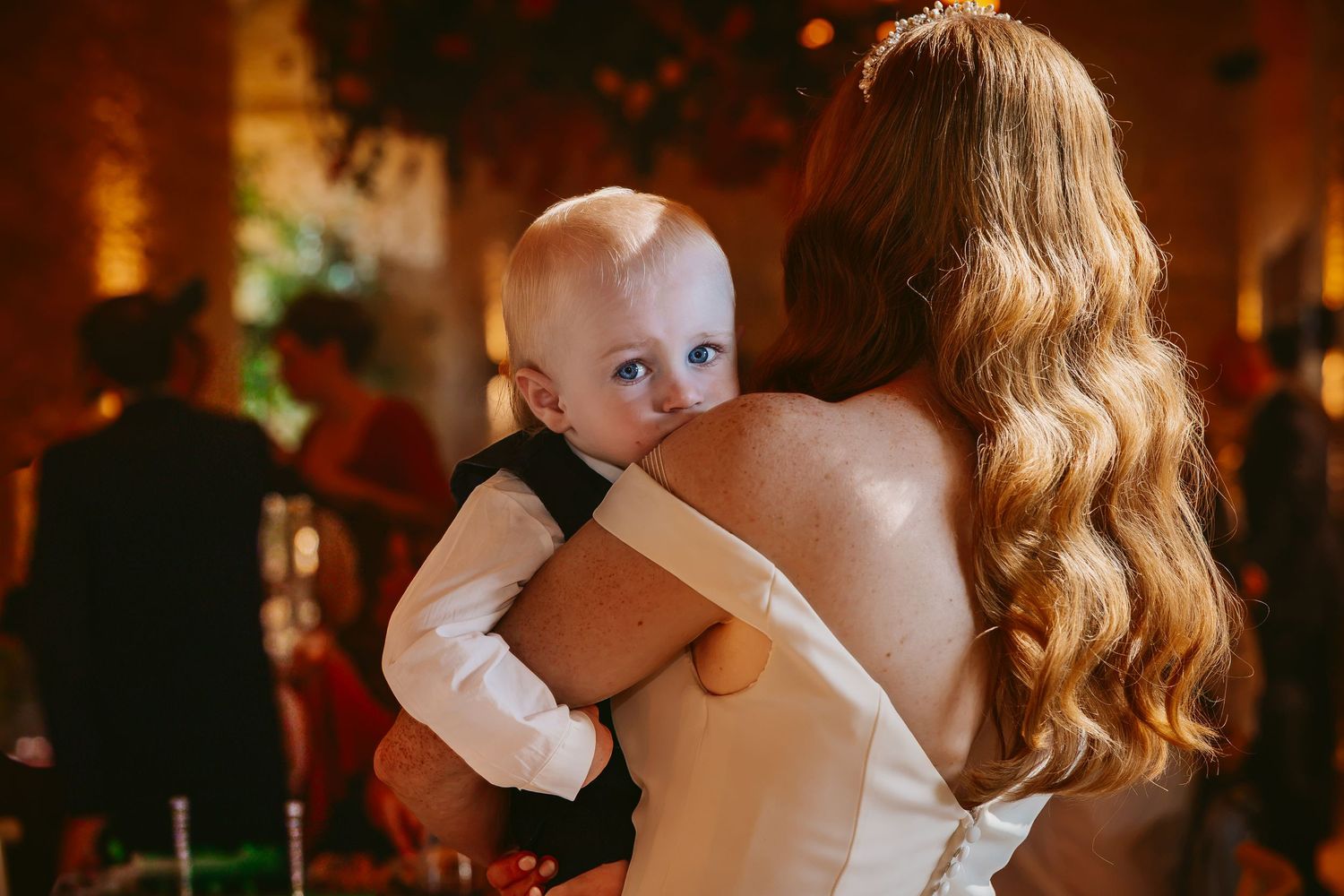 Bride in a white gown with long wavy red hair holds a curious baby in a warmly lit wedding reception venue.