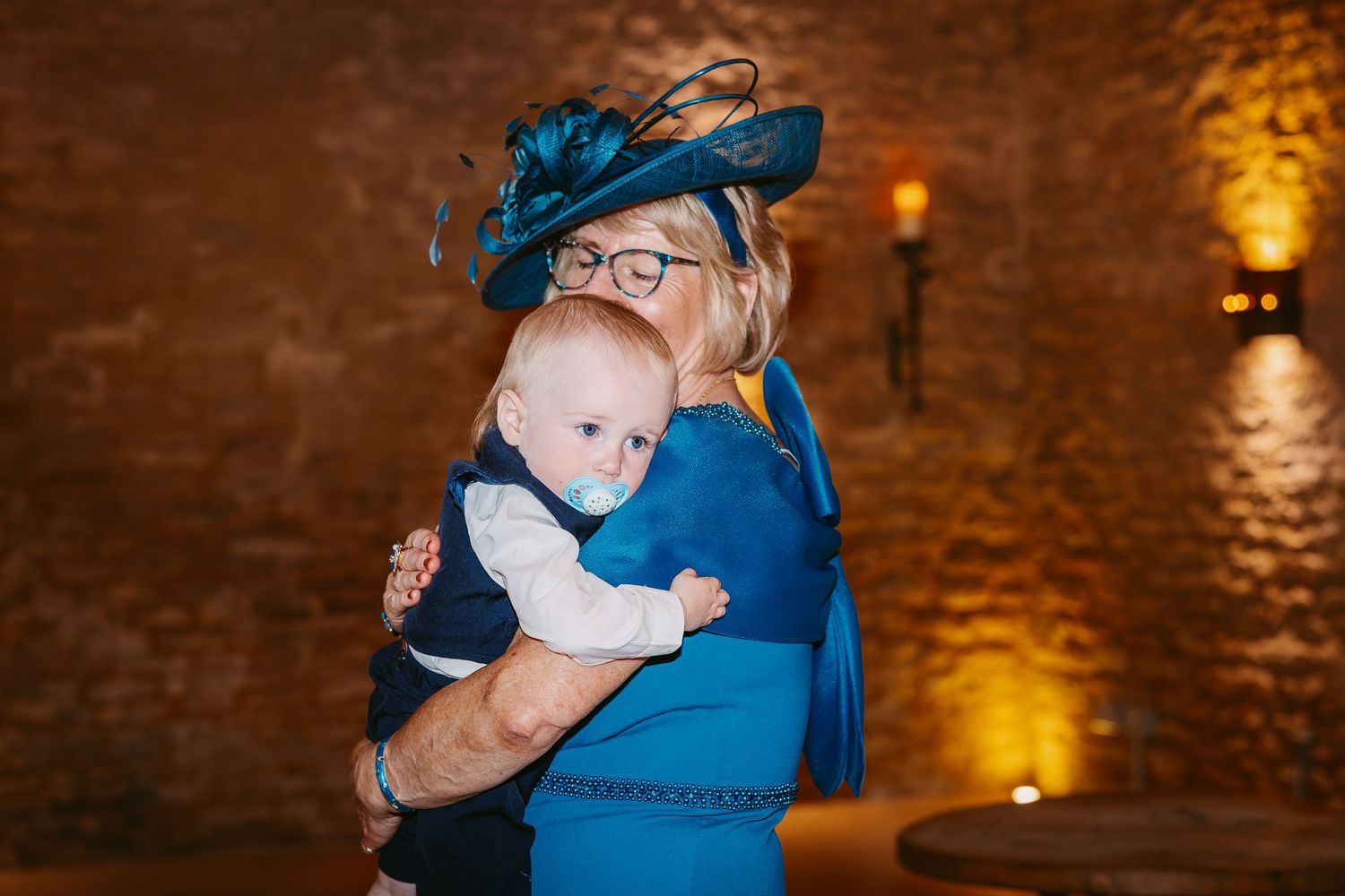 A woman in a blue dress and navy fascinator hat holds a baby wearing a pacifier at a candlelit wedding reception.
