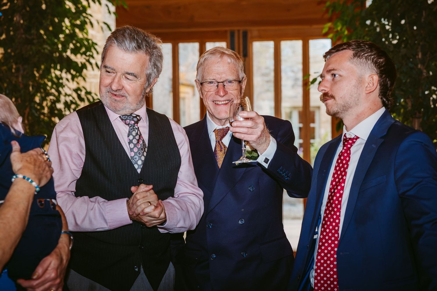 Three smiling male wedding guests in suits and ties chat and hold drinks during a celebratory reception.