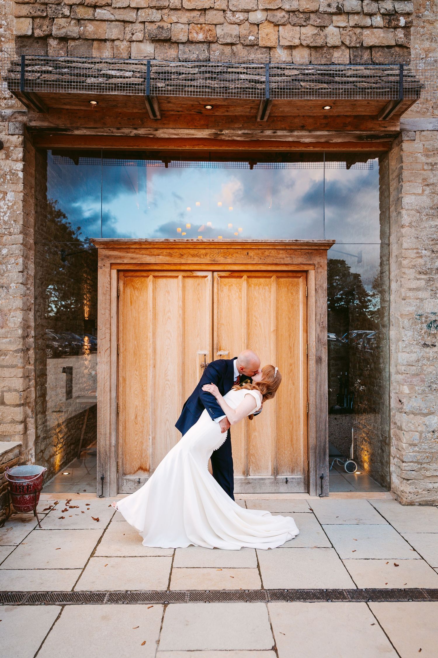 Groom dips the bride in a dramatic wedding kiss in front of tall wooden barn doors and a rustic stone building.