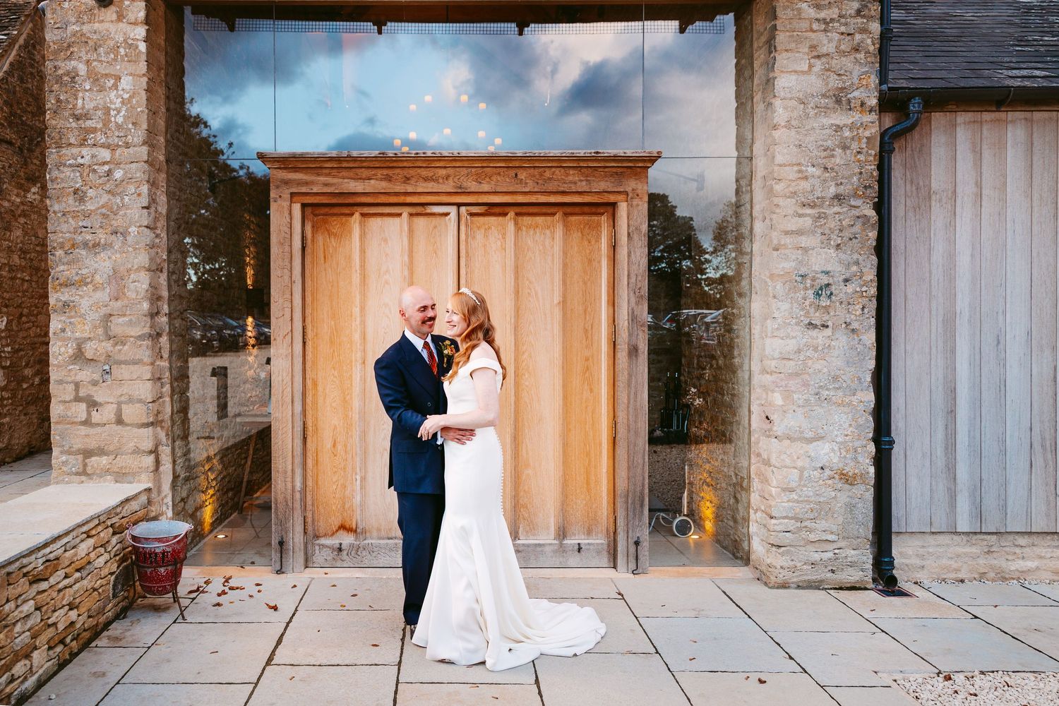 Bride in a fitted white gown and groom in a navy suit embrace on a stone patio in front of large wooden barn doors.