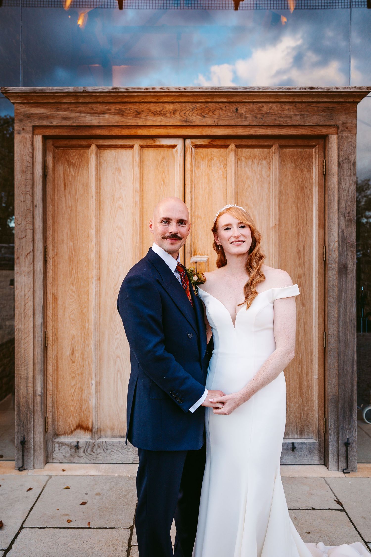 Smiling bride in an off-the-shoulder white gown and groom in a navy suit pose together in front of a large rustic wooden door.