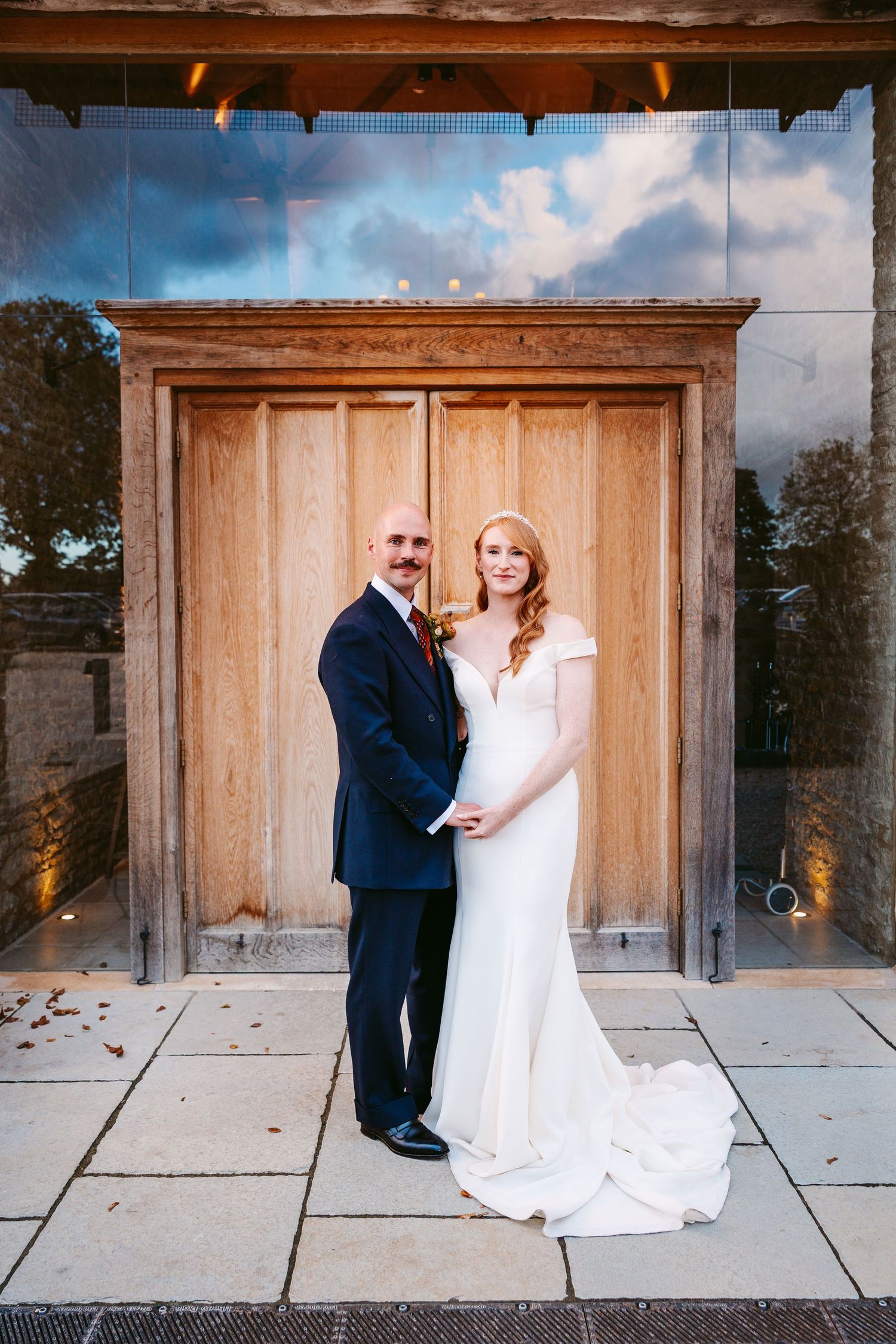 A bride in a white gown and a groom in a navy suit hold hands and pose in front of a large wooden door at their wedding venue.