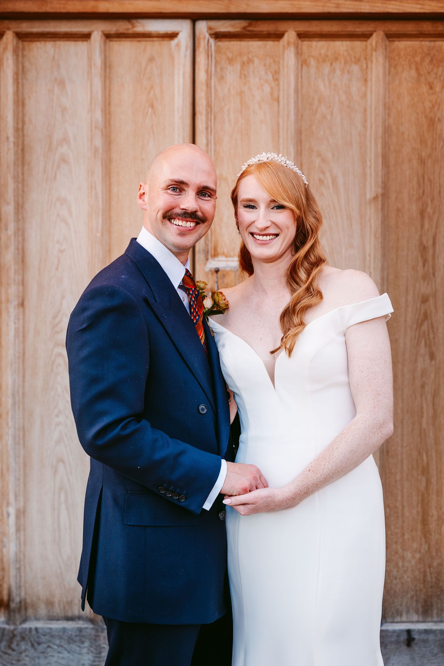 Smiling bride with a floral tiara and groom with a moustache pose together in front of a large wooden door.