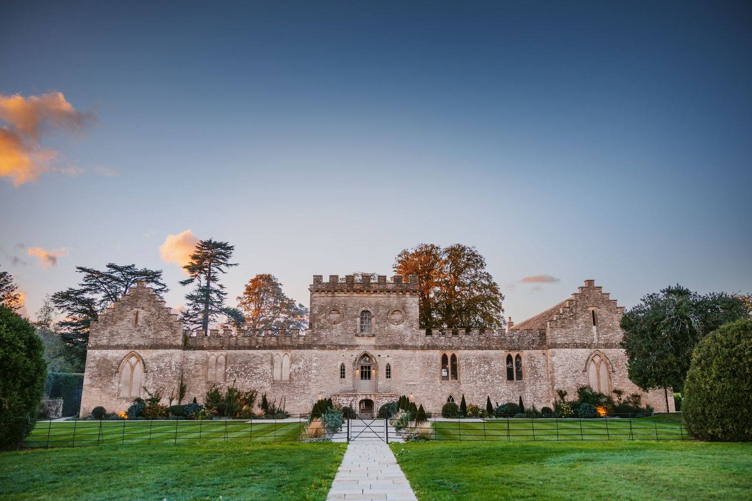 Historic stone castle venue with battlements stands behind a manicured lawn and pathway at sunset, ideal for weddings.