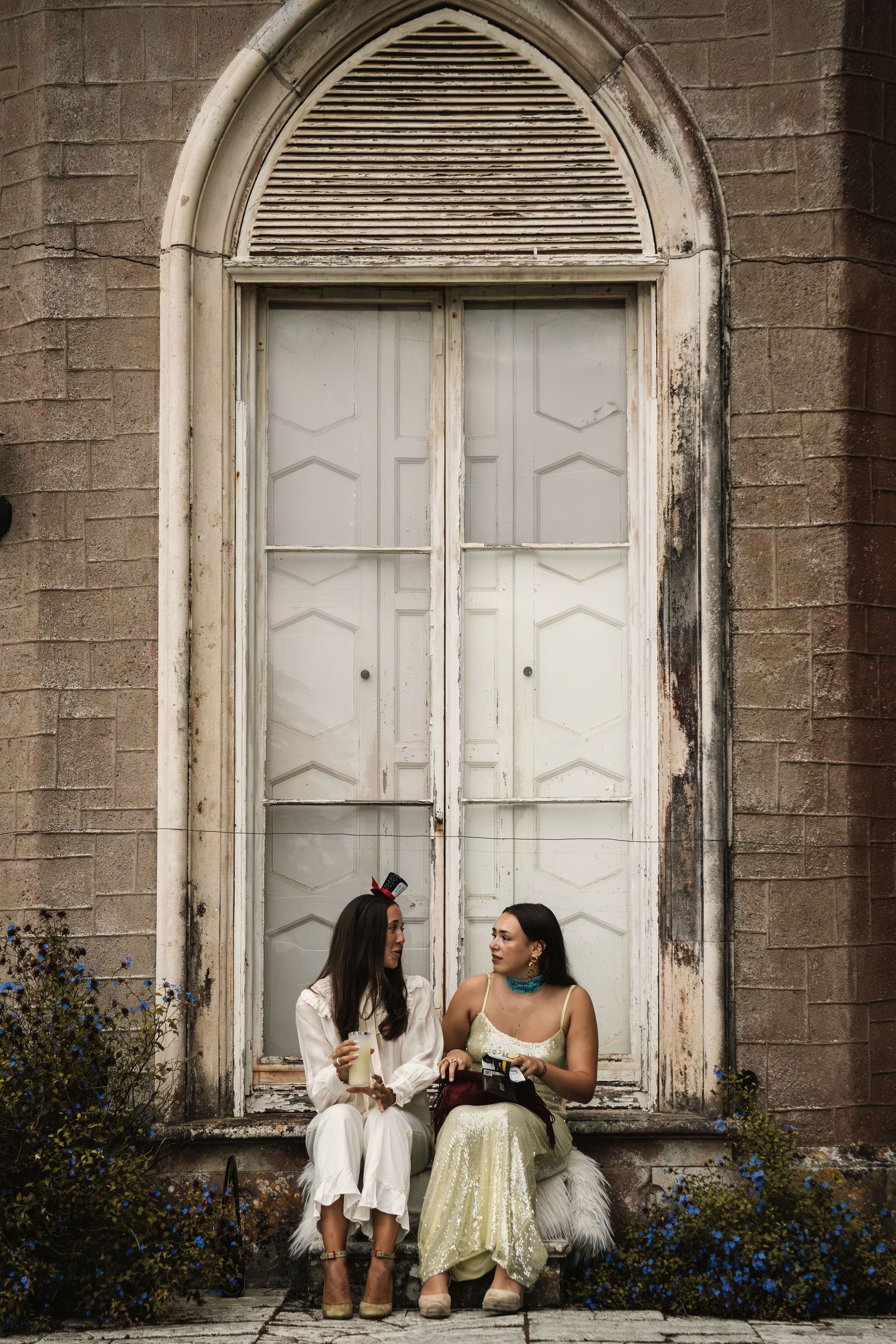 Two women in bohemian outfits sitting and chatting in front of a rustic arched white door on stone steps.