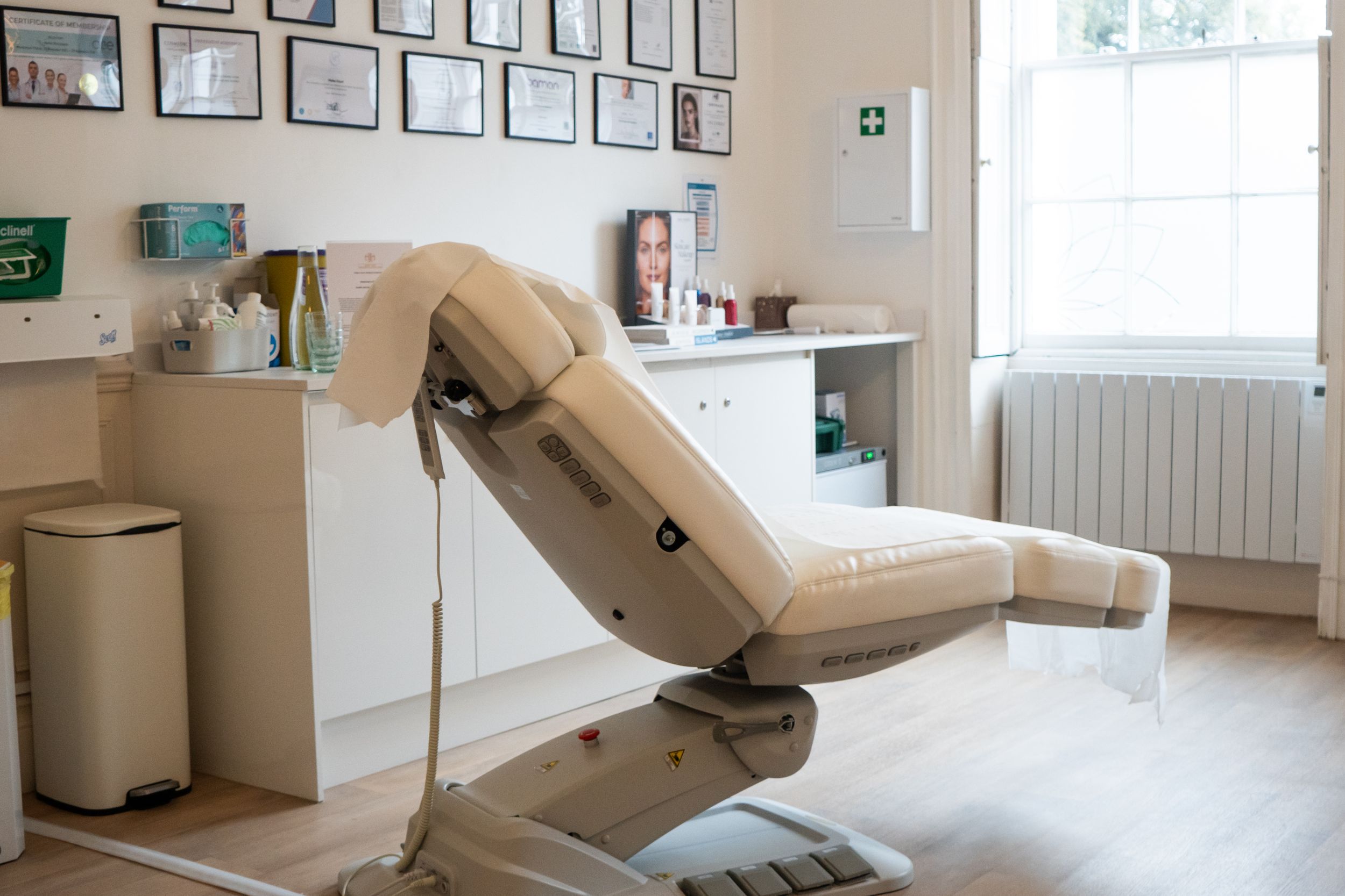 A modern medical reclining chair in a bright clinical treatment room with framed certificates on the wall.