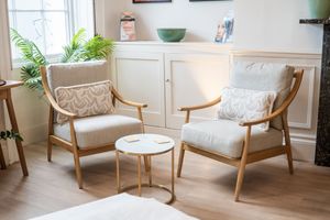Two mid-century modern wooden armchairs with beige cushions flank a small marble side table in a bright, cozy living room.