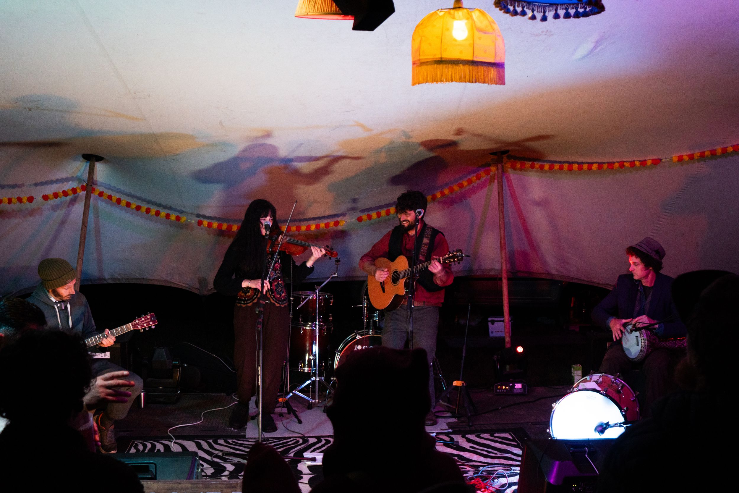 Band performing live music in a colorful tent venue with warm lighting, guitars, and an intimate audience gathering.
