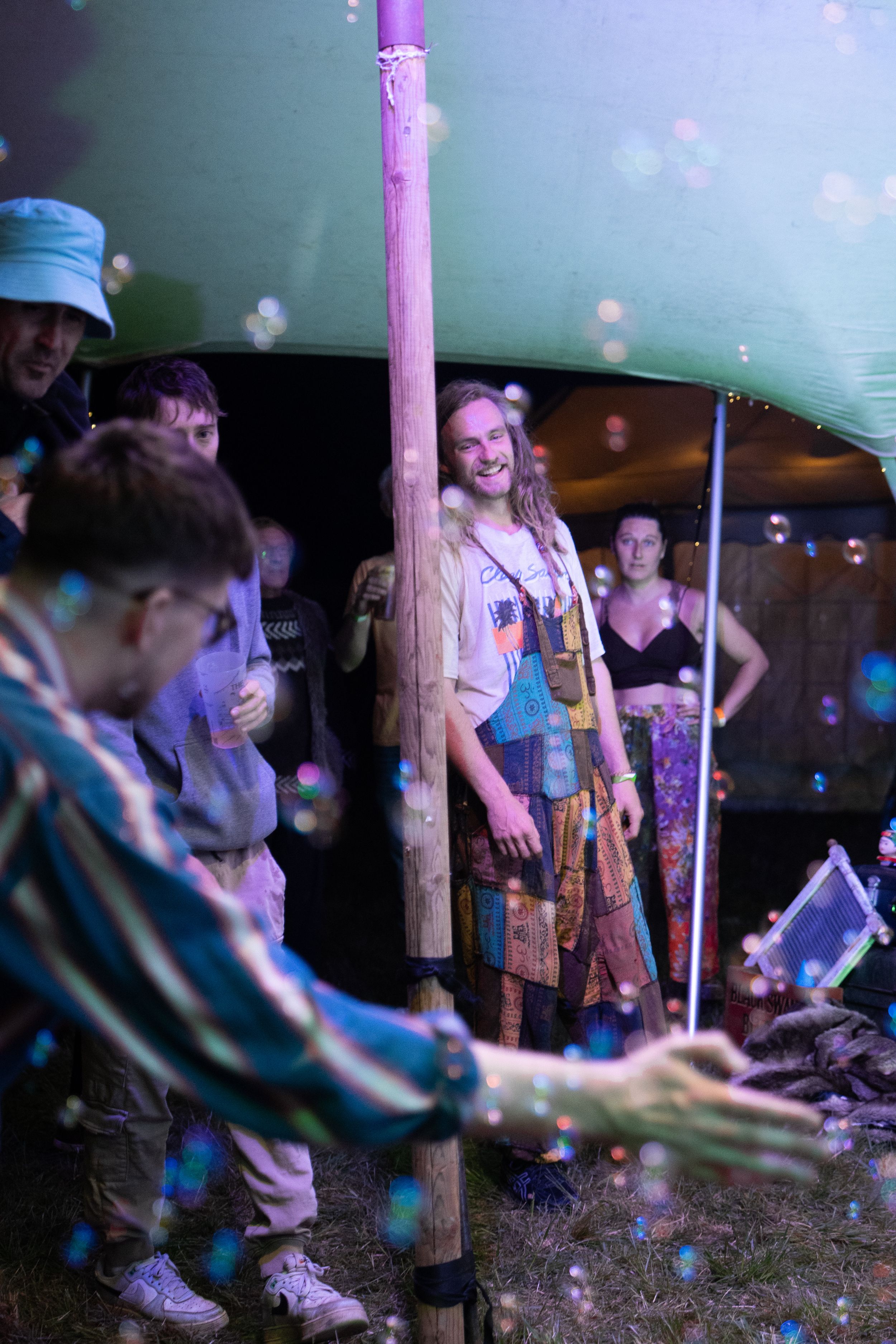 People enjoying a festival under a green tent with colorful bubbles floating in the air, wearing bohemian clothing.