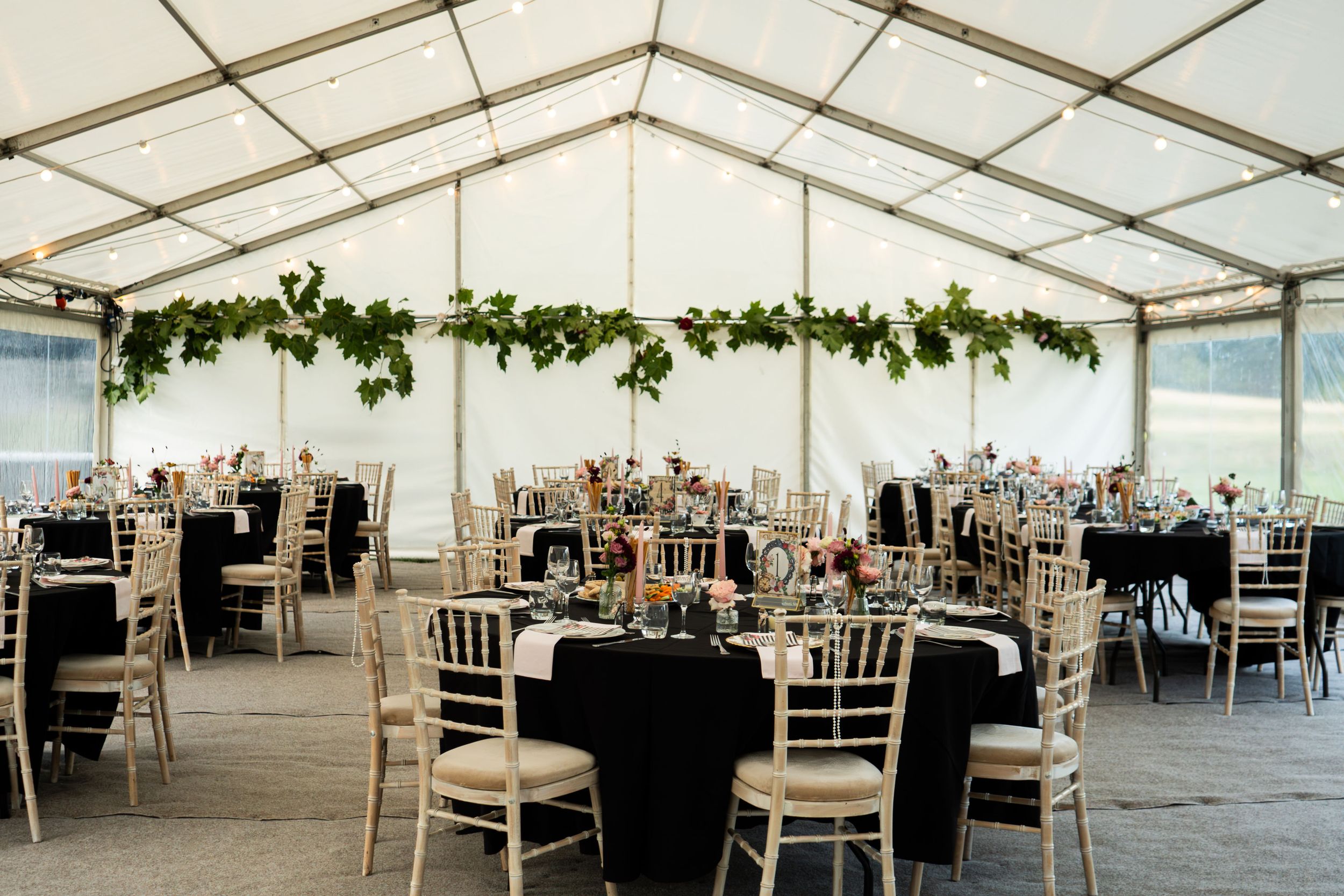 Elegant wedding reception tent with black tablecloths, gold chiavari chairs, and lush green garland overhead.