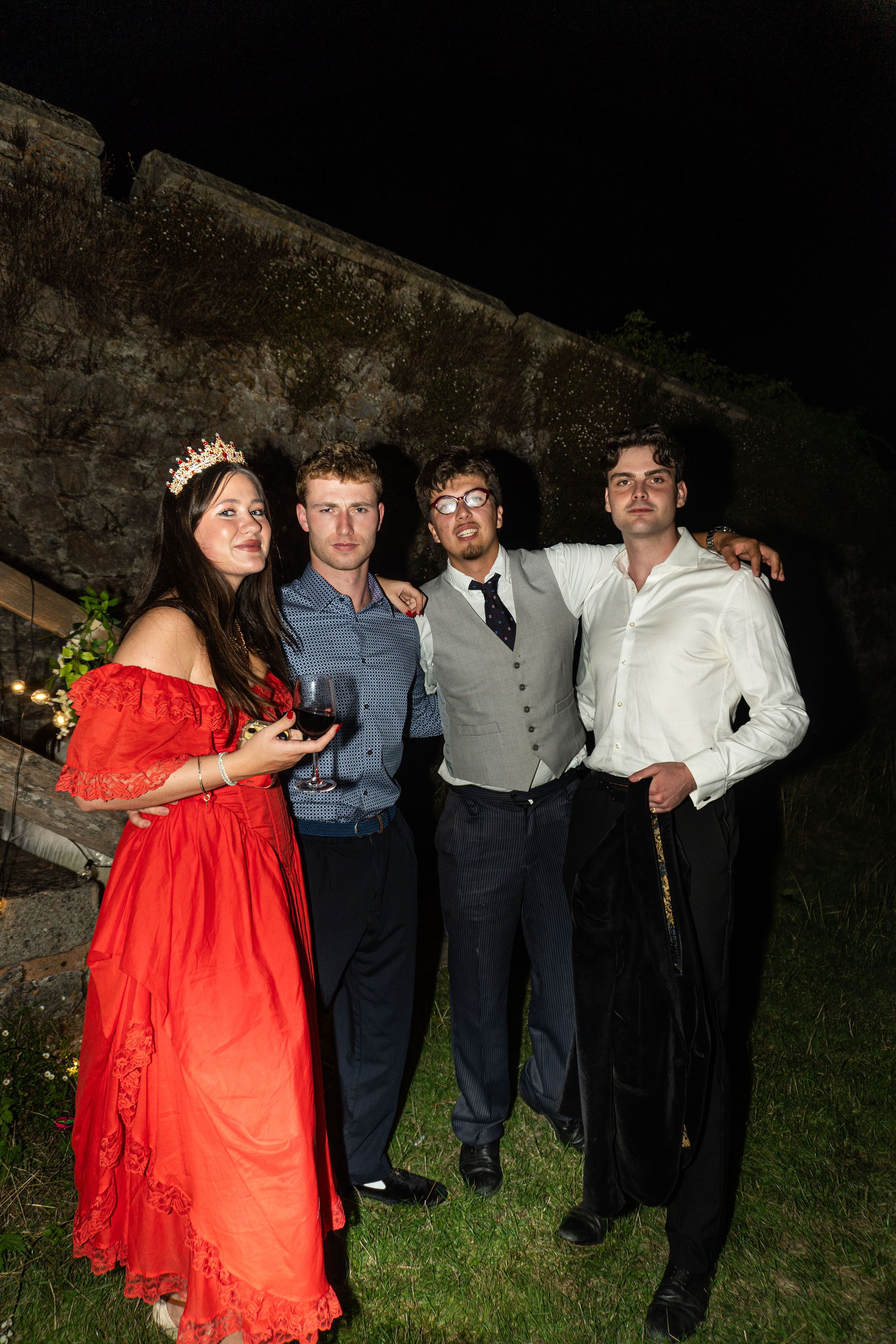 Four elegantly dressed young adults posing outdoors at night, woman in red dress wearing tiara holding wine glass.
