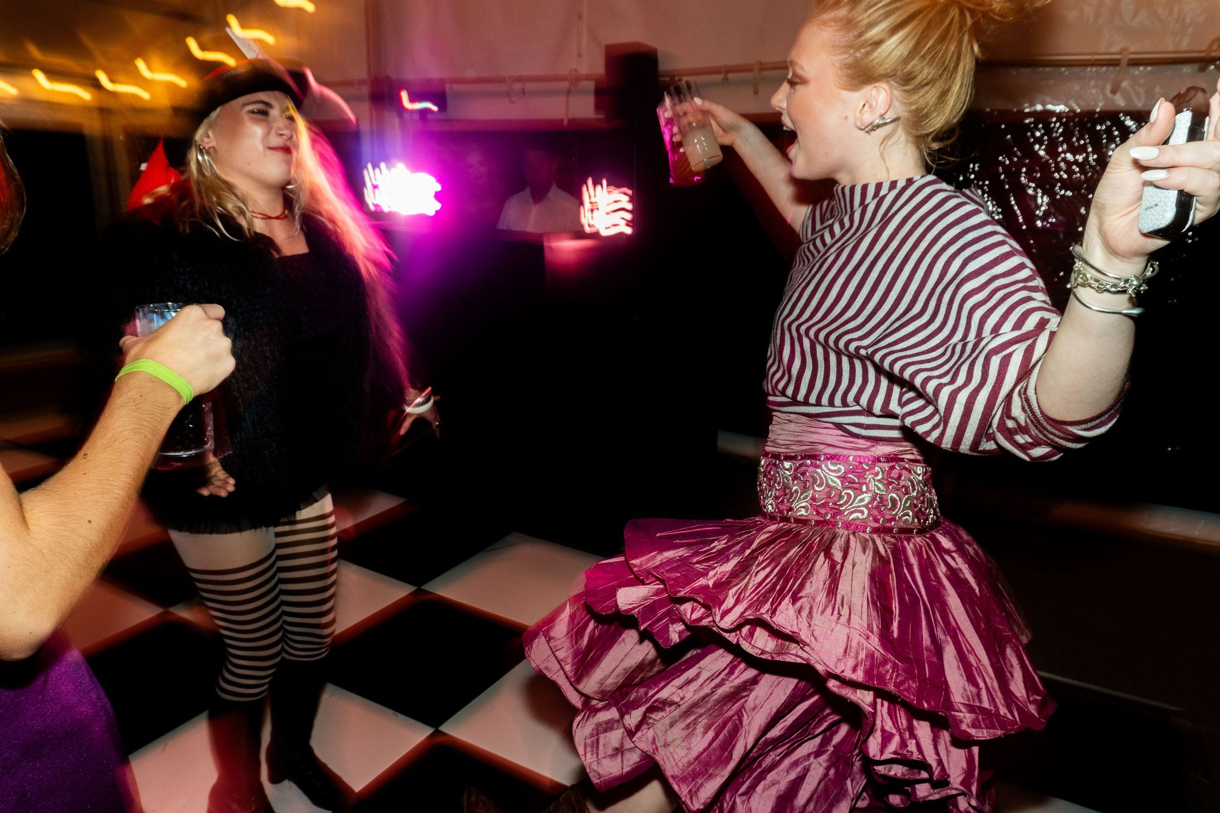 Two women dancing at a party, one in a pink tutu skirt and striped top, the other in fishnet stockings and pink hair.