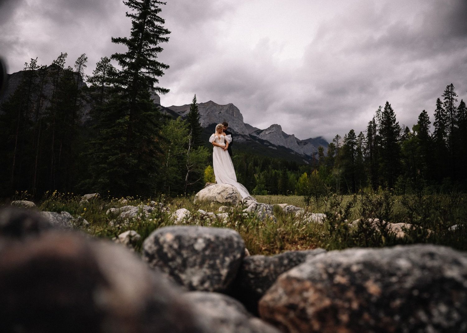 Bride and groom on a boulder in a mountain meadow under moody clouds, Canmore wedding photographer
