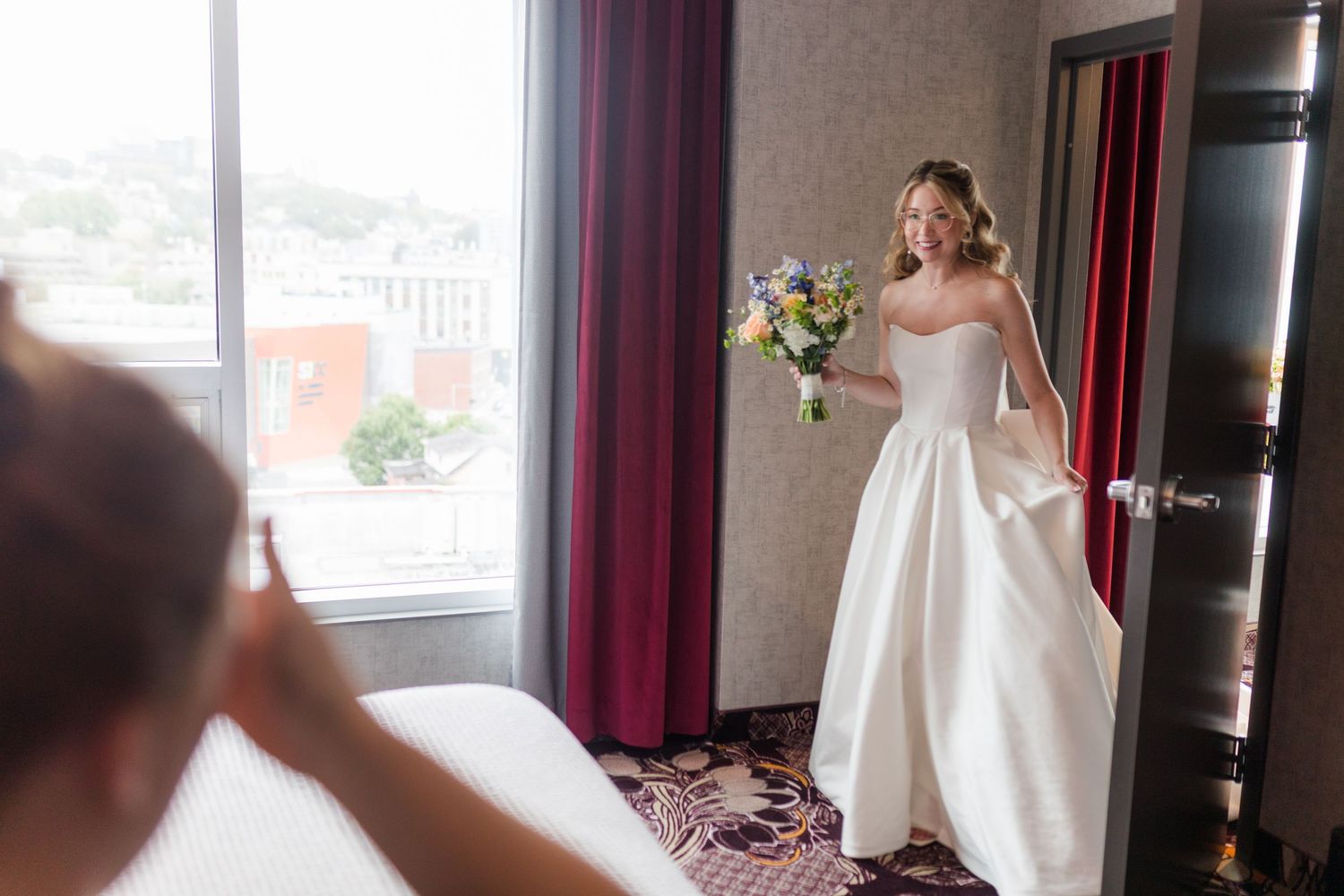 Bride in strapless white gown holding colorful bouquet poses in hotel room with red curtains and city view.