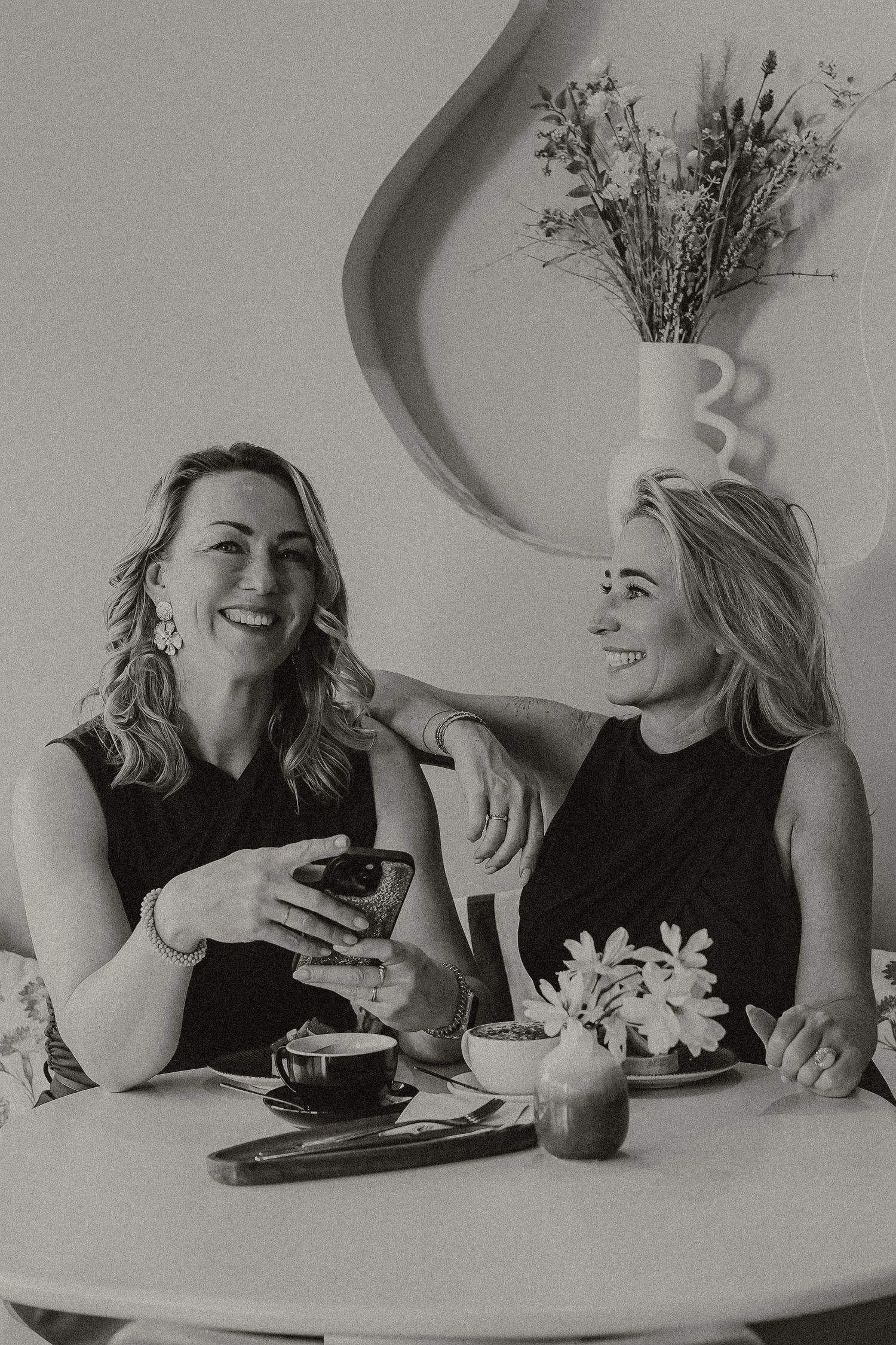 Two women laughing and enjoying coffee together at a cozy cafe booth with elegant decor.