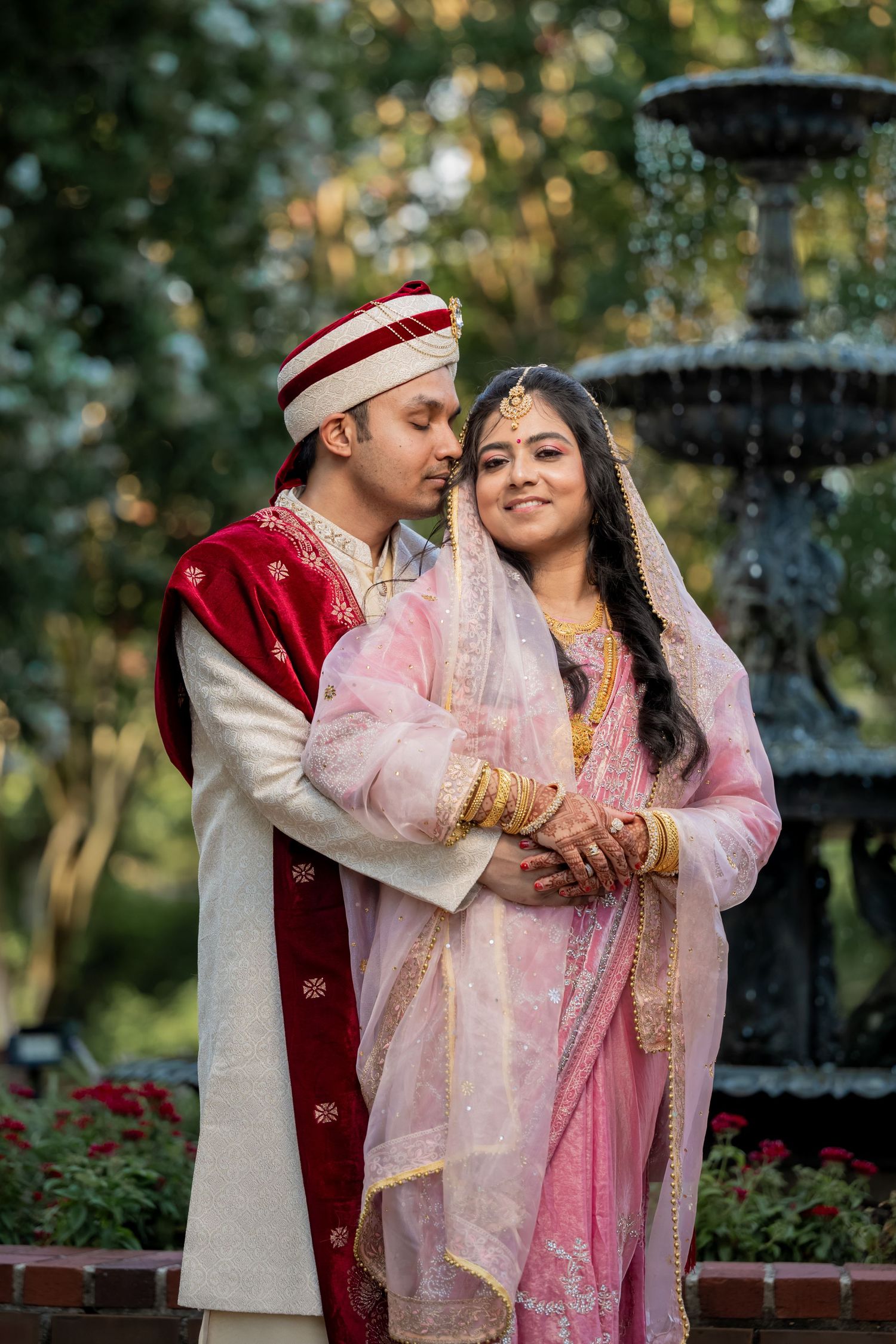 Couple in traditional South Asian wedding attire embracing in front of a stone fountain - by our Collierville wedding photographer