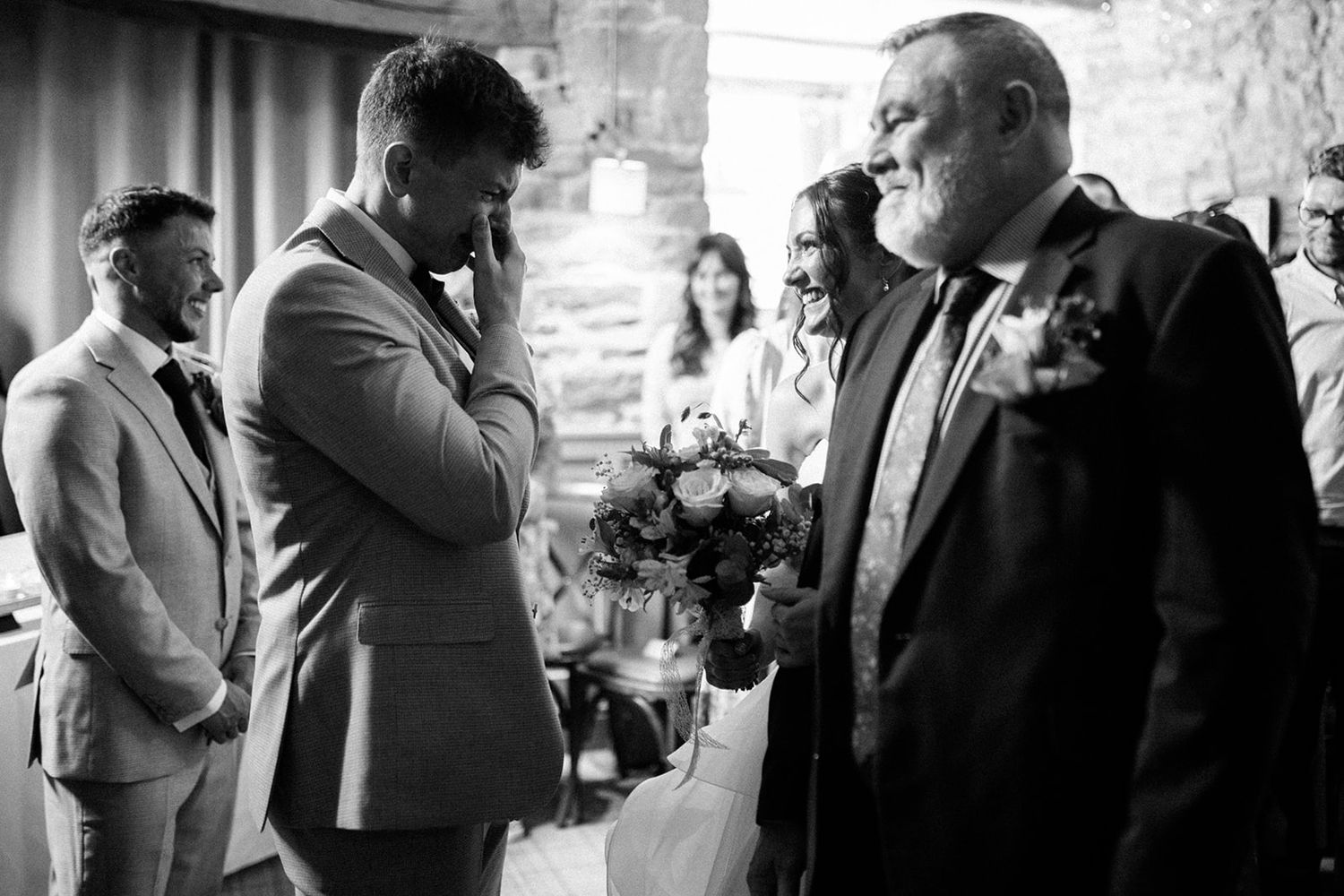 Emotional groom in a gray suit wipes his eyes as a bride with a bouquet approaches during a black and white wedding ceremony.