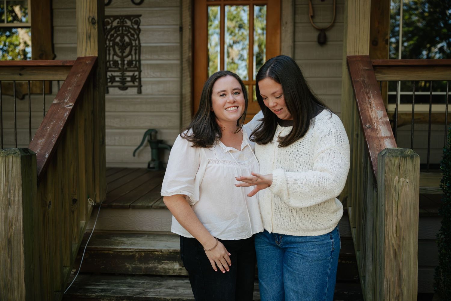 Two smiling women embrace outdoors as one shows off her sparkling engagement ring on her raised hand in front of a rustic wooden building.