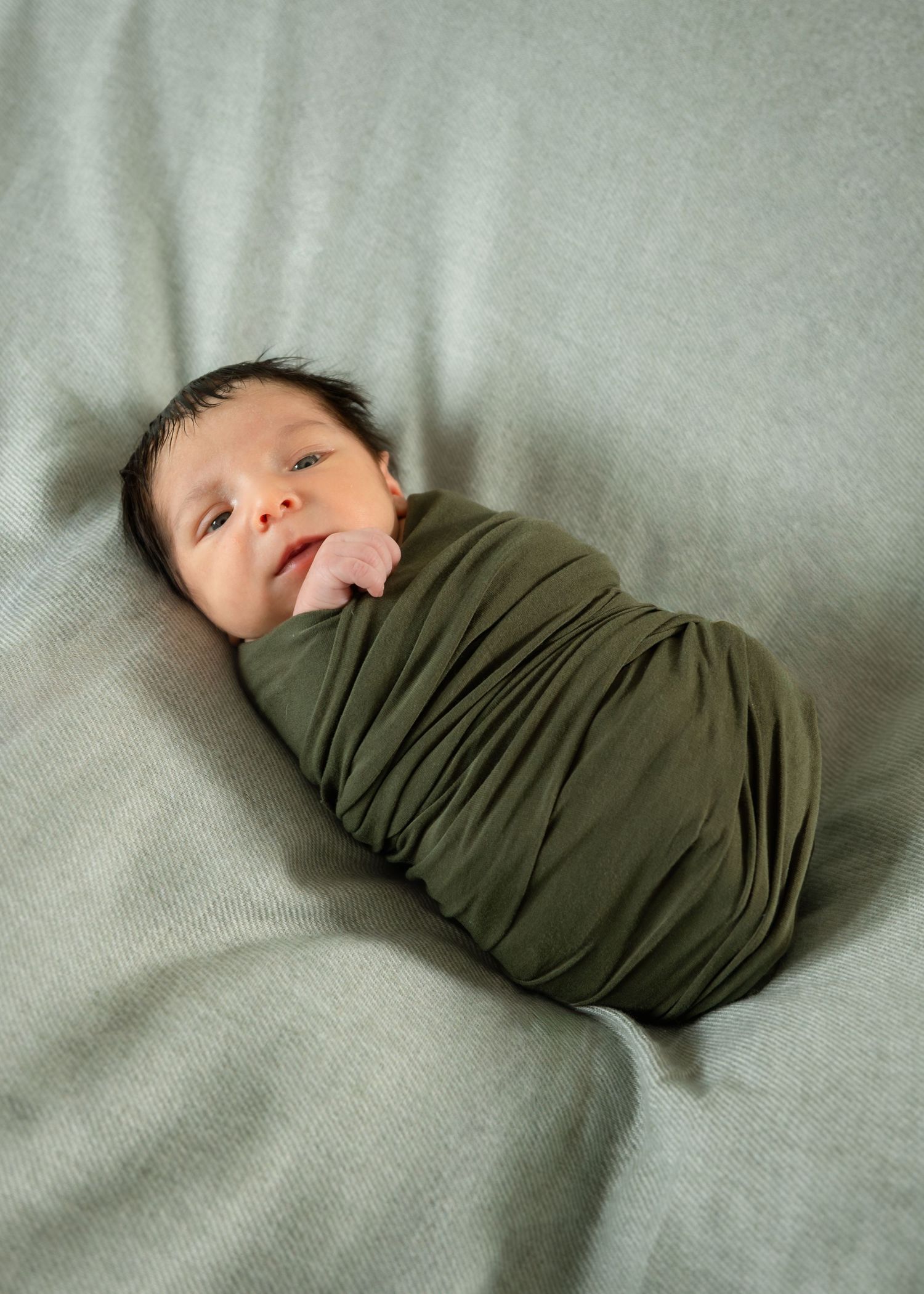 Newborn baby swaddled in olive green wrap, lying on soft gray blanket with dark hair and tongue out.
