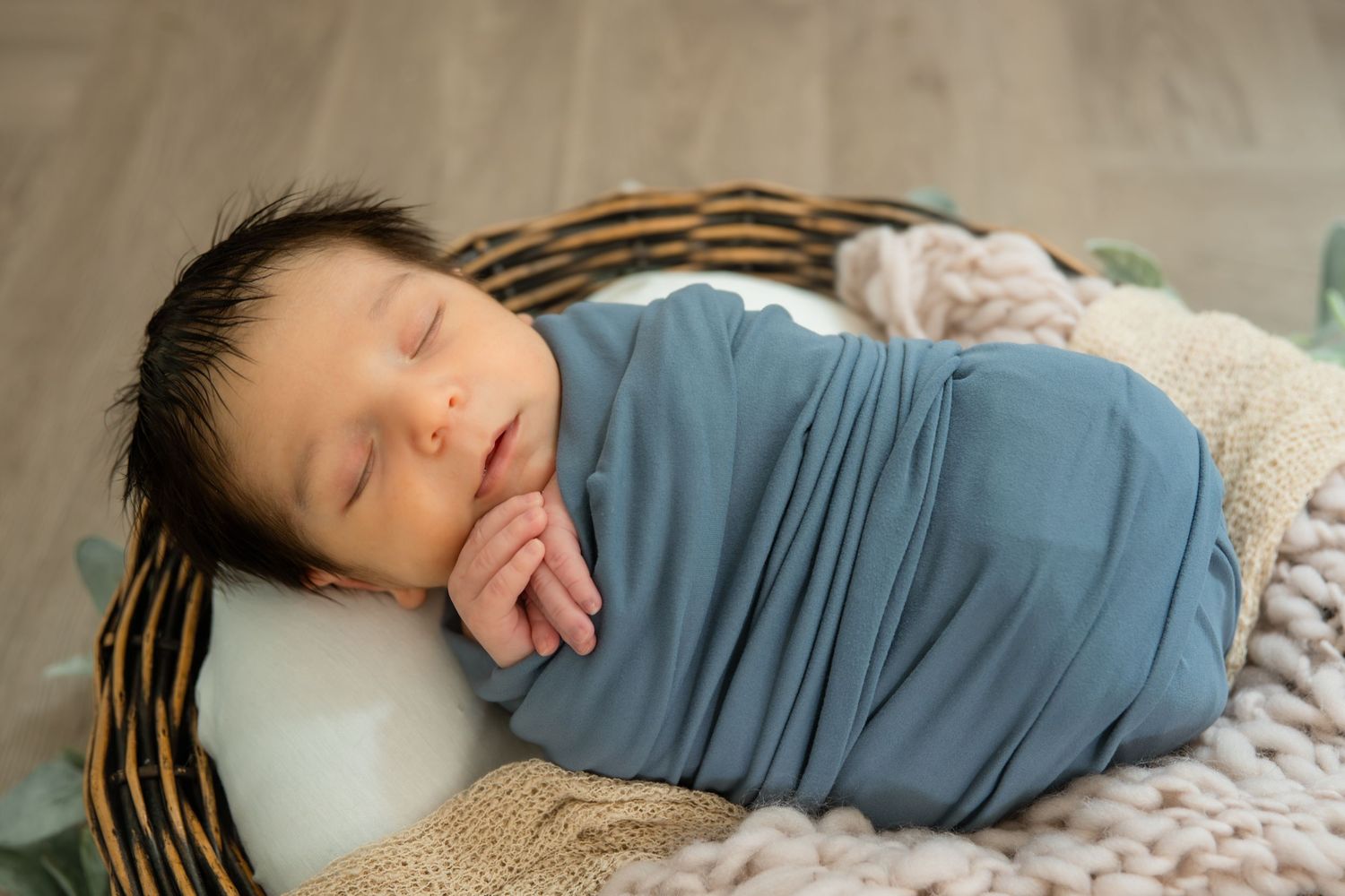 Newborn baby swaddled in blue wrap sleeping peacefully in a wicker basket with soft blankets.