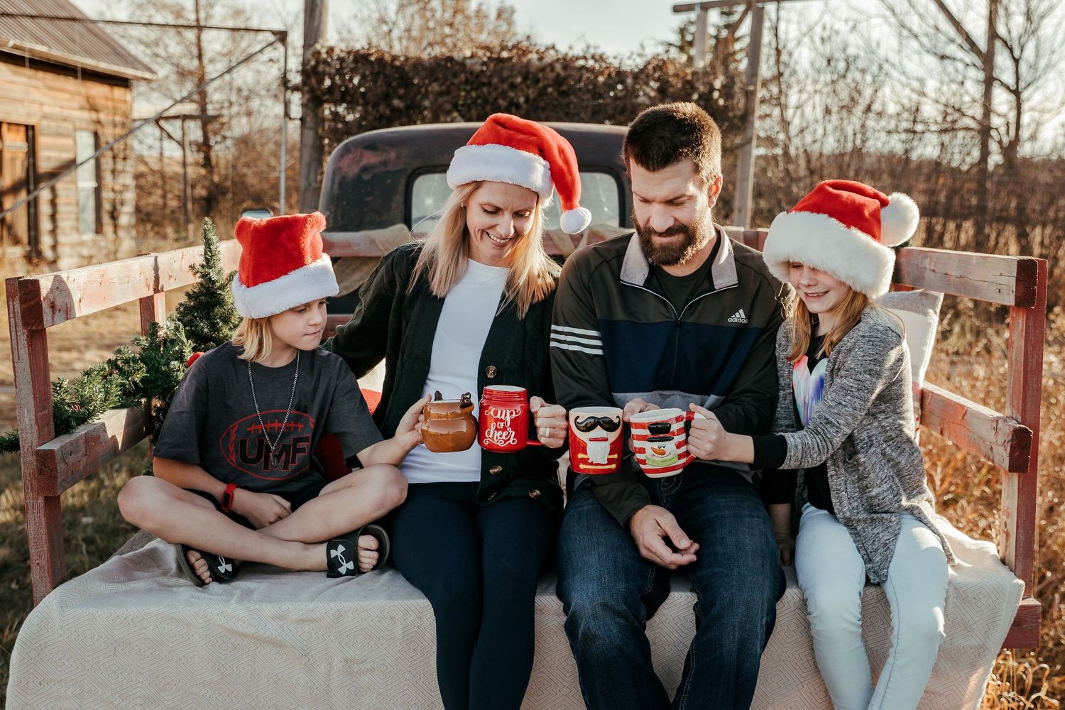 Smiling family in Santa hats sits on a rustic bench near a vintage truck, holding red mugs during a cozy outdoor Christmas photo session.
