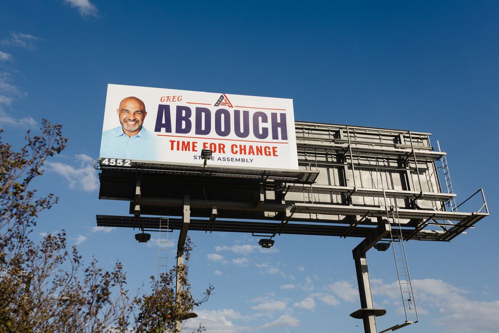 Large roadside billboard reading Abdouch, Time for Change, shown against a bright blue sky with tree branches in the foreground.