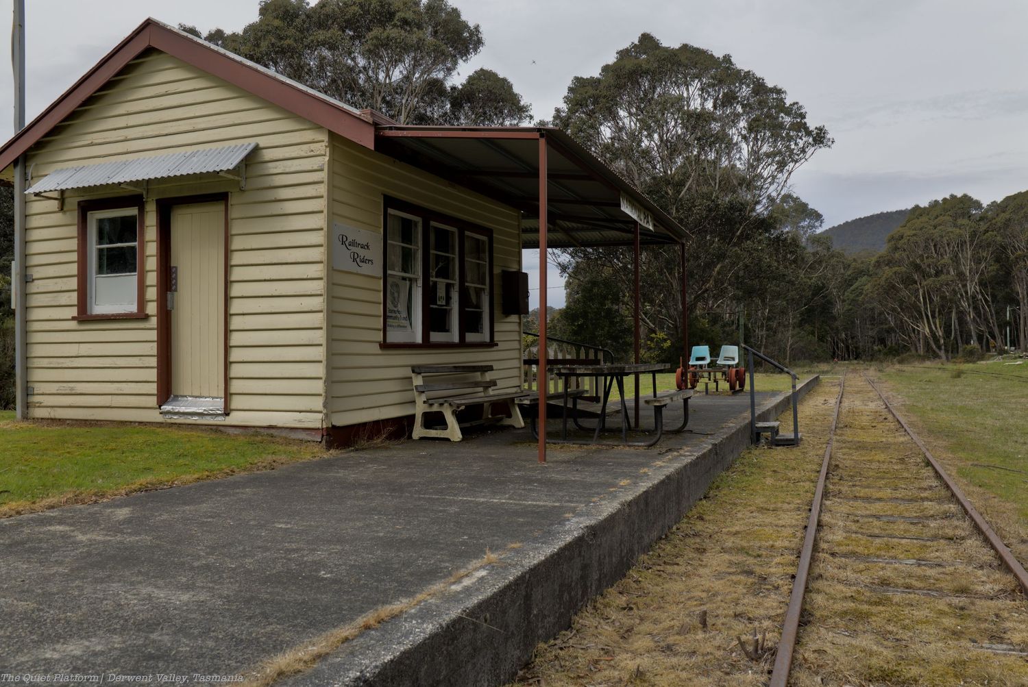 Small rustic wooden railway station with a corrugated roof, picnic tables, and a concrete platform beside overgrown tracks in a bushland set
