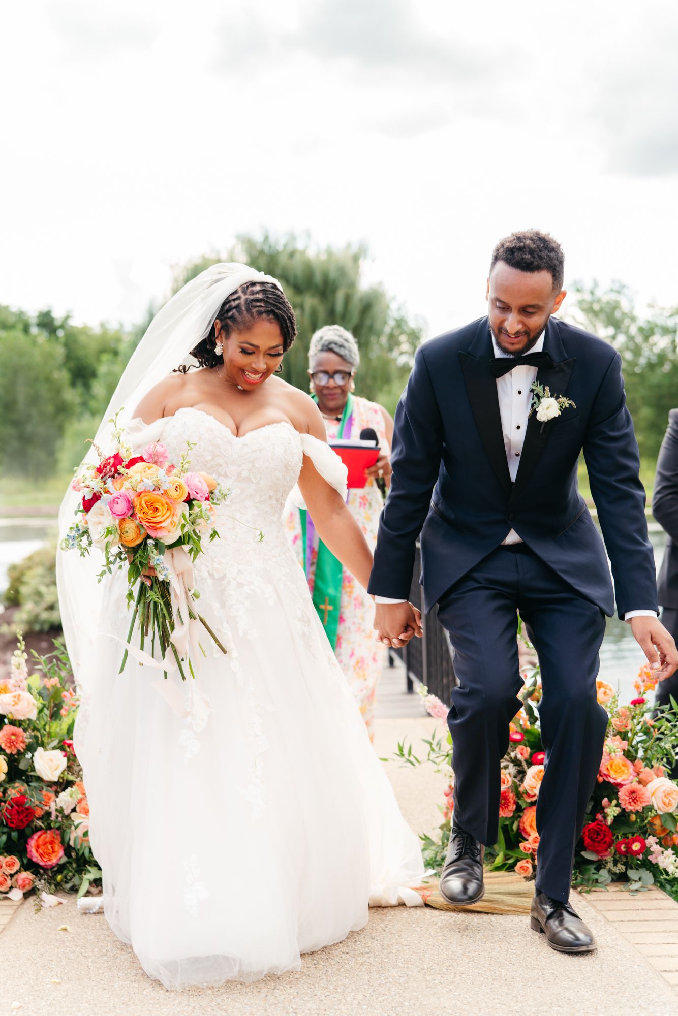 Smiling bride in a white off-the-shoulder gown and veil holds hands with her groom in a navy tuxedo during an outdoor wedding ceremony.