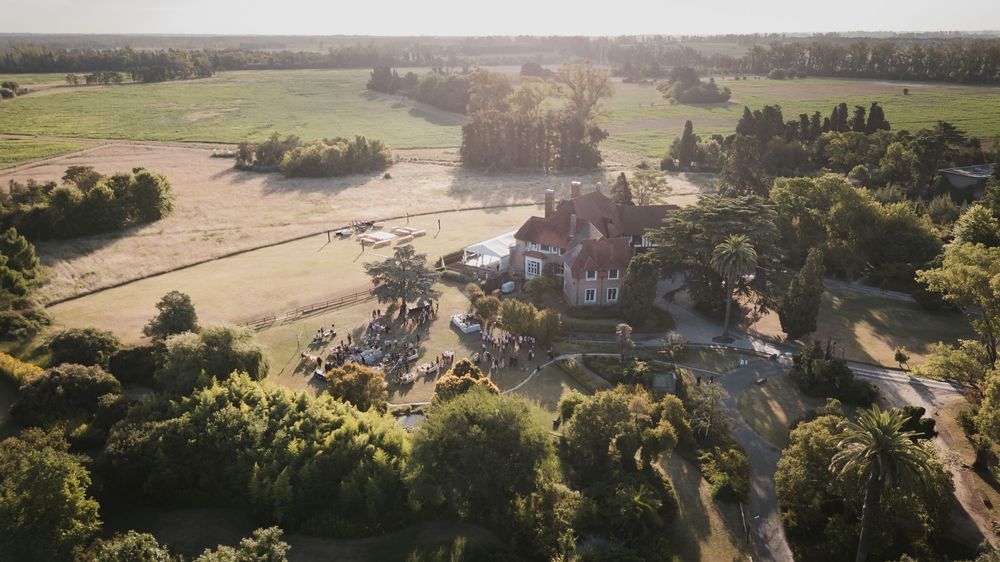 Aerial view of a historic estate with a red-roofed manor house surrounded by lush trees, gardens, and open green fields at sunset.