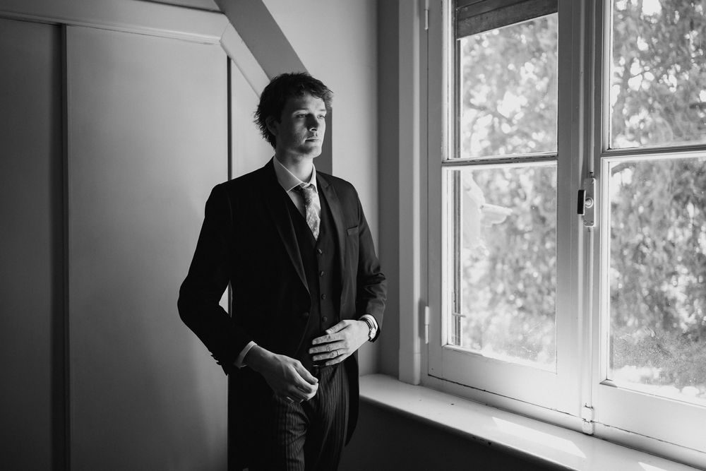 Black and white portrait of a man in a dark three-piece suit and tie standing beside a large window, gazing outside in soft natural light.
